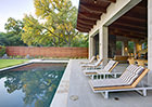 Modern square pool surrounded by patio with large stone fireplace, ceiling with wooden beams, and custom hardwood vertical fence